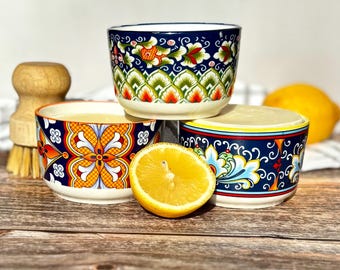 May include: Three ceramic bowls with intricate, colorful patterns in shades of blue, orange, green, and white. Each bowl contains a solid, light-colored substance. A halved lemon sits in the foreground, and a wooden scrub brush is visible nearby.