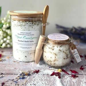 May include: Two glass jars of herbal bath salts with wooden scoops. One jar is labeled "Mint Eucalyptus" and the other "Lavender." The jars have wooden lids and are tied with twine. The background includes scattered flower petals and sprigs.