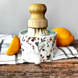 May include: A white ceramic citrus juicer with a floral design and a wooden reamer. A halved orange and a whole lemon are placed nearby. The juicer is on a striped towel on a wooden surface. The juicer is filled with juice.