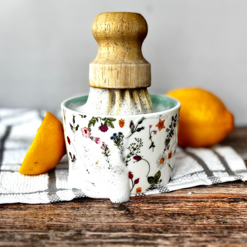 May include: A white ceramic citrus juicer with a floral design and a wooden reamer. A halved orange and a whole lemon are placed nearby. The juicer is on a striped towel on a wooden surface. The juicer is filled with juice.