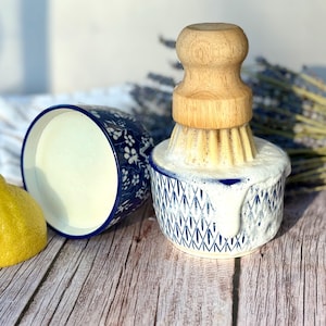 May include: A round ceramic bowl with a blue and white geometric pattern containing white soap suds and a wooden dish brush with light-coloured bristles. A second blue and white floral bowl and a yellow lemon are positioned nearby.