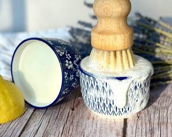 May include: A round ceramic dish with a blue and white geometric pattern, containing white soap suds and a wooden dish brush with light-colored bristles. A second blue and white floral ceramic dish and a yellow lemon are positioned nearby.