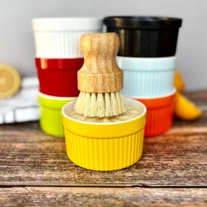 May include: A yellow ramekin with a wooden-handled brush, filled with a light-colored substance. Behind it are stacks of colorful ramekins in white, red, green, light blue, orange, and black. The ramekins are on a wooden surface.