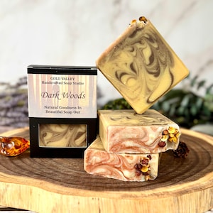 May include: Three rectangular bars of soap with a marbled cream and brown swirl pattern. One bar is inside a black box labeled Gold Valley Handcrafted Soap Studio Dark Woods. The other two bars are stacked, featuring dried flower accents.