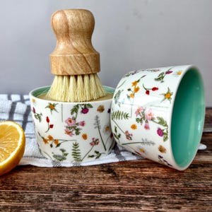 May include: A white ceramic dish with a floral pattern, a light green interior, and a wooden-handled brush. The dish is decorated with colorful flowers and greenery. A half lemon is visible on the left.