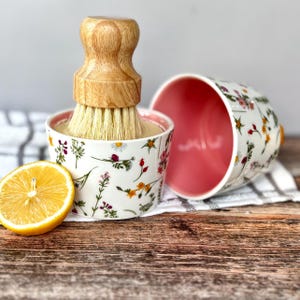 May include: A white ceramic bowl with a floral pattern and a pink interior, next to a matching bowl. A wooden-handled brush sits inside the bowl. A lemon half is on a wooden surface.