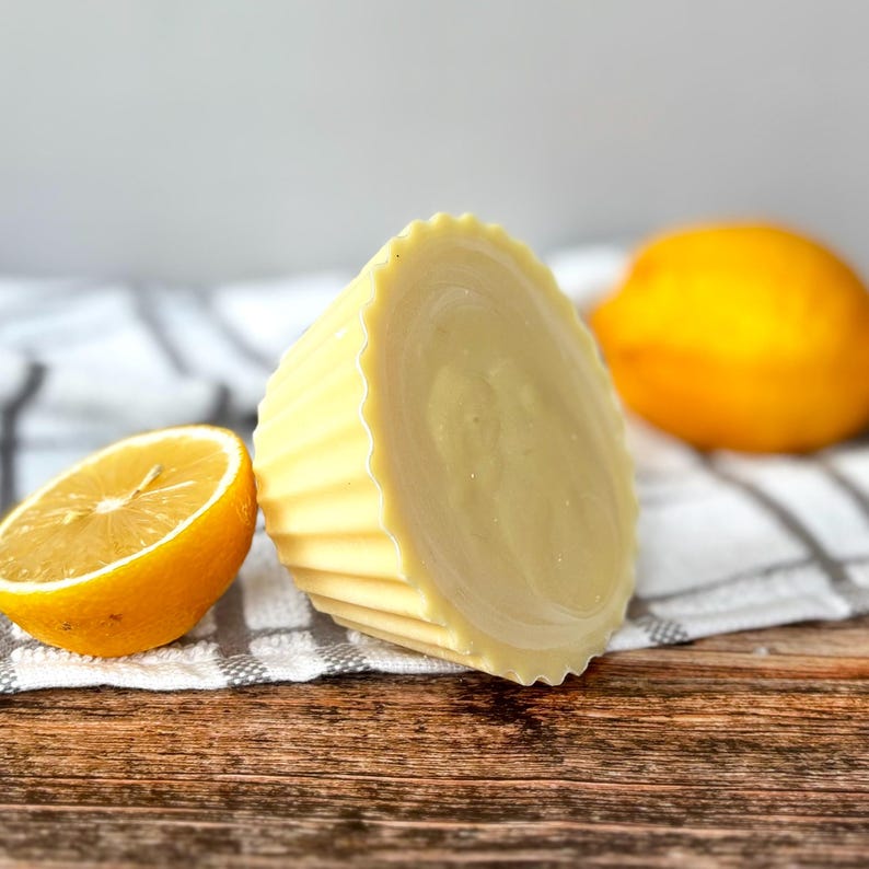 May include: A pale yellow, fluted soap bar, cut in half, beside a lemon half and a whole lemon. The soap has a circular pattern on the cut surface. The items are on a wooden surface with a black and white striped cloth.
