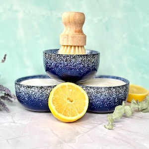 May include: Three dark blue ceramic bowls with a white speckled pattern, arranged in a pyramid. A wooden dish brush with light-colored bristles rests inside the top bowl. A bright yellow lemon slice is placed in front of the bottom two bowls.