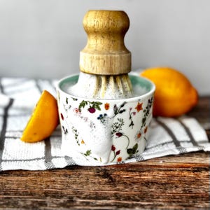 May include: A white ceramic citrus juicer with a wooden reamer, adorned with colourful floral patterns. A halved orange and a whole lemon are placed nearby. The juicer is on a wooden surface.