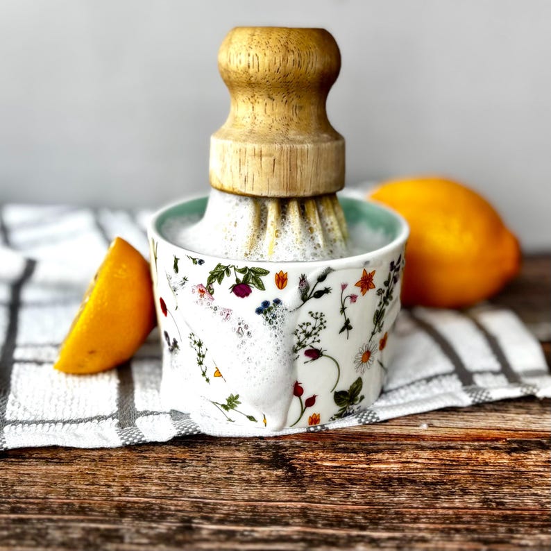 May include: A white ceramic citrus juicer with a wooden reamer, adorned with colourful floral patterns. A halved orange and a whole lemon are placed nearby. The juicer is on a wooden surface.