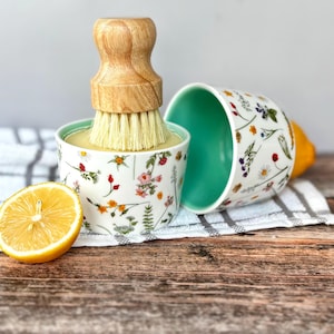 May include: A white ceramic citrus juicer with a wooden handle and brush, adorned with a floral design. A halved lemon and an additional bowl are on a striped towel and wooden surface. The juicer is for squeezing citrus fruits.