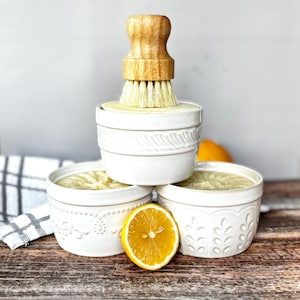 May include: Three white ceramic bowls with decorative patterns, filled with a light-coloured substance. A wooden-handled brush is placed on top. A halved lemon sits in front of the bowls on a wooden surface.