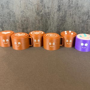 May include: A collection of six miniature coffee mugs with smiling faces. Five are brown with different latte art designs on top, and one is purple with a colorful sprinkle design. Each mug has a handle.