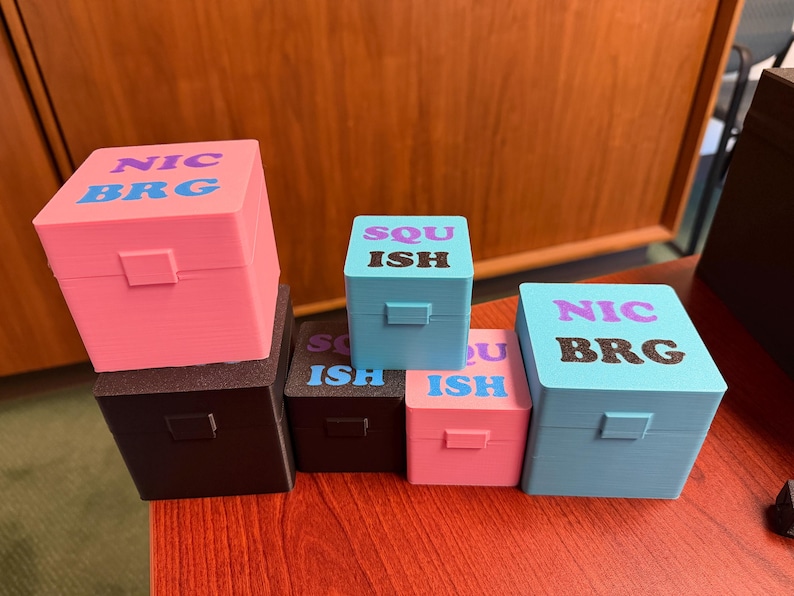 May include: A collection of colorful, small, square boxes with hinged lids. The boxes are in shades of pink, blue, and black. Each box has text on the top, including "NIC BRG" and "SQU ISH". The boxes are arranged on a red surface.
