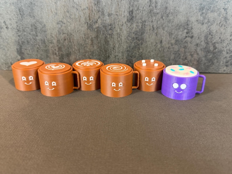 May include: A collection of six miniature coffee mugs with smiling faces. Five are brown with different latte art designs on top, and one is purple with a colorful sprinkle design. Each mug has a handle.