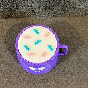 May include: A purple 3D-printed mug with a white top decorated with blue and pink sprinkles. The mug has a handle and a smiling face design on the front.