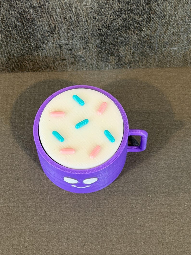 May include: A purple 3D-printed mug with a white top decorated with blue and pink sprinkles. The mug has a handle and a smiling face design on the front.
