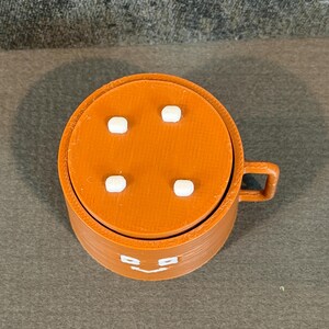 May include: A 3D-printed orange mug with a lid featuring four white square accents. The mug has a handle and a smiling face design on the front. The mug is made of plastic.