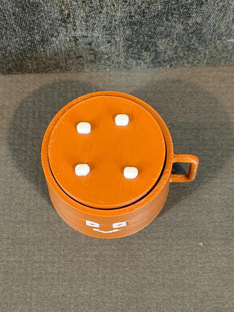 May include: A 3D-printed orange mug with a lid featuring four white square accents. The mug has a handle and a smiling face design on the front. The mug is made of plastic.
