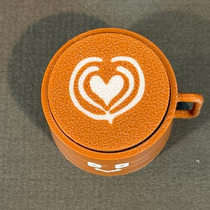 May include: An orange coffee cup with a white heart-shaped design on top. The cup has a small handle and a smiling face design on the side. The cup is made of a textured material.