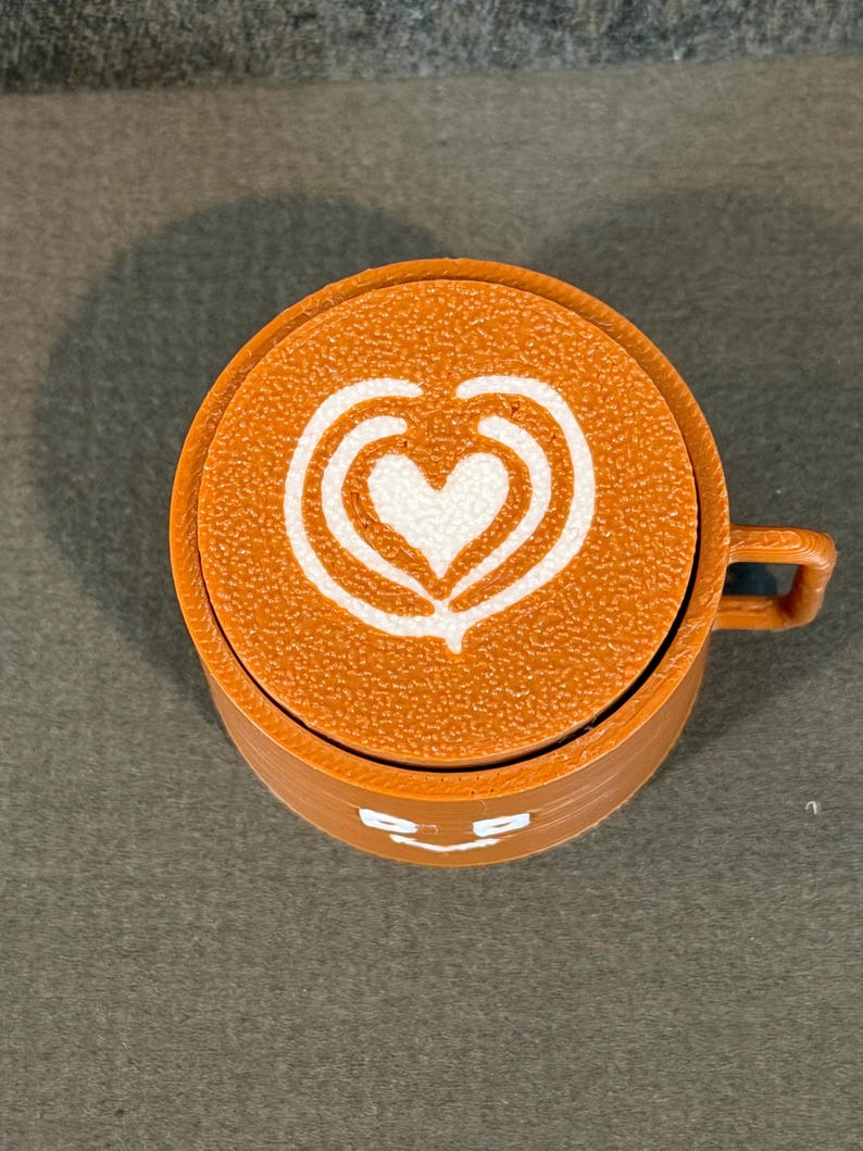 May include: An orange coffee cup with a white heart-shaped design on top. The cup has a small handle and a smiling face design on the side. The cup is made of a textured material.