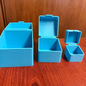 May include: Three open, light blue, square-shaped boxes with hinged lids are displayed on a dark wood surface. The boxes vary in size, with the largest on the left and the smallest on the right. The lids are open, revealing the interior of the boxes.