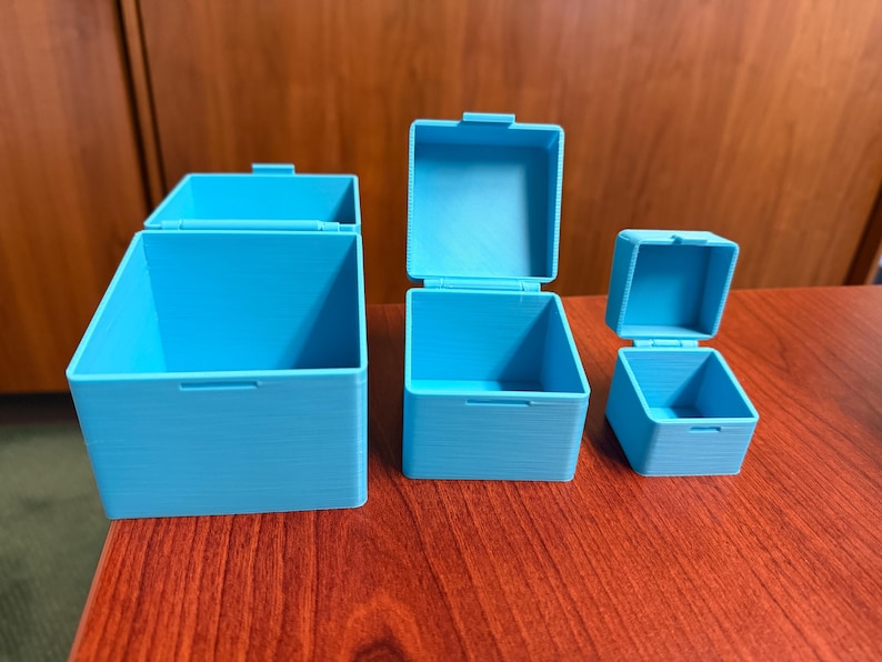 May include: Three open, light blue, square-shaped boxes with hinged lids are displayed on a dark wood surface. The boxes vary in size, with the largest on the left and the smallest on the right. The lids are open, revealing the interior of the boxes.