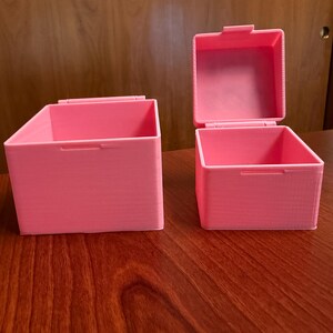 May include: Two pink storage boxes with hinged lids sit on a wooden surface. One box is open, revealing the interior. The boxes are rectangular with a textured surface and a handle on the front.