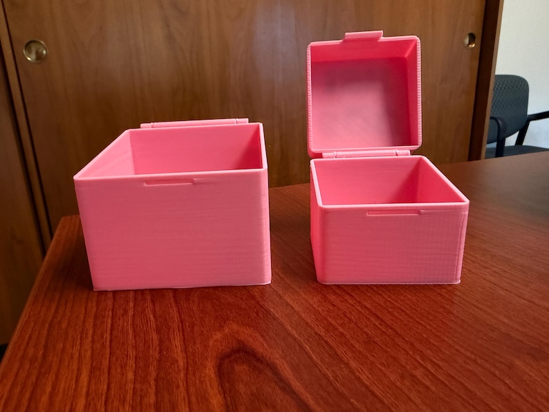May include: Two pink storage boxes with hinged lids sit on a wooden surface. One box is open, revealing the interior. The boxes are rectangular with a textured surface and a handle on the front.