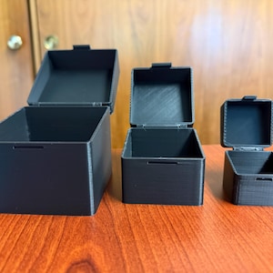 May include: Three black square boxes of varying sizes with hinged lids, arranged on a wooden surface. The boxes are open, revealing their empty interiors. The lids are slightly ajar, and the boxes have a textured appearance.