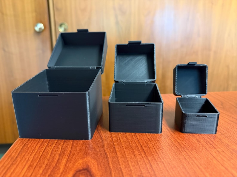 May include: Three black square boxes of varying sizes with hinged lids, arranged on a wooden surface. The boxes are open, revealing their empty interiors. The lids are slightly ajar, and the boxes have a textured appearance.