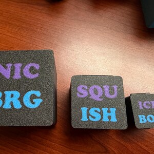 May include: Three black foam cubes of varying sizes with text. The largest cube has "NIC" in purple and "BRG" in blue. The medium cube reads "SQU" in purple and "ISH" in blue. The smallest cube says "ICE BOX" in purple.