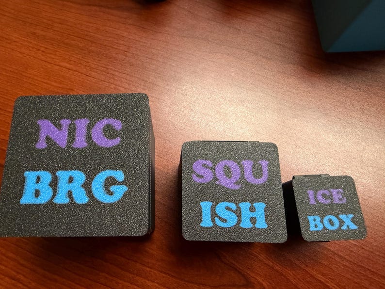 May include: Three black foam cubes of varying sizes with text. The largest cube has "NIC" in purple and "BRG" in blue. The medium cube reads "SQU" in purple and "ISH" in blue. The smallest cube says "ICE BOX" in purple.