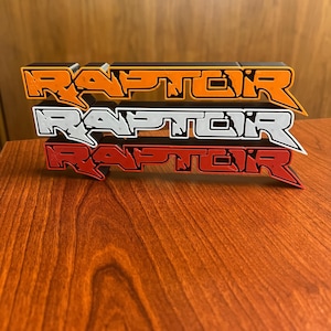 May include: Three "RAPTOR" logo emblems in orange, white, and red, stacked on a wooden surface. The emblems have a 3D effect with black outlines, and the orange emblem is at the top.