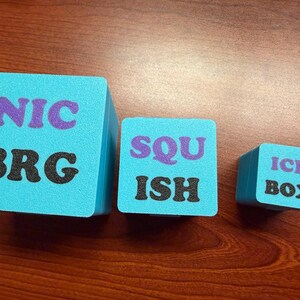 May include: Three turquoise foam cubes with black and purple text. The largest cube reads "NIC BRG", the middle cube says "SQU ISH", and the smallest cube says "ICE BOX". The cubes are arranged on a wooden surface.