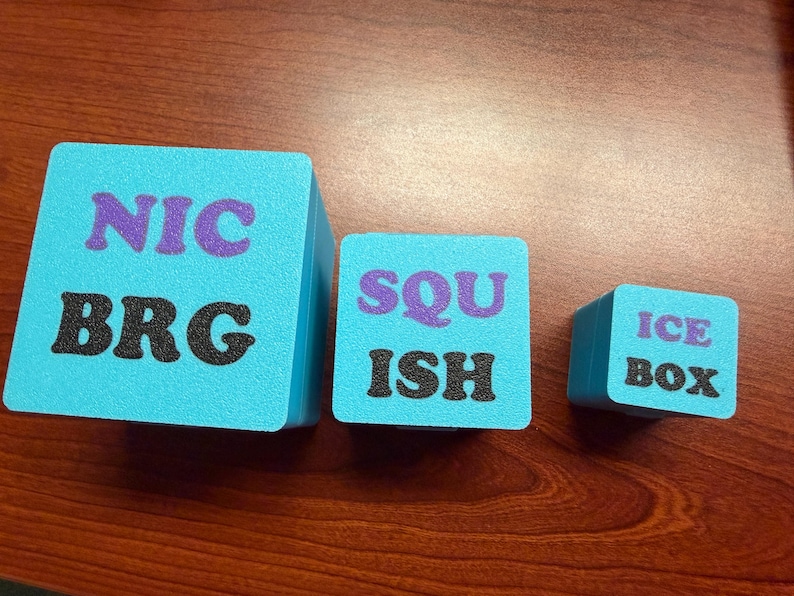 May include: Three turquoise foam cubes with black and purple text. The largest cube reads "NIC BRG", the middle cube says "SQU ISH", and the smallest cube says "ICE BOX". The cubes are arranged on a wooden surface.