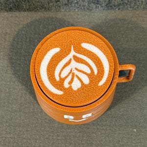 May include: A brown coffee cup with a white latte art design on top. The cup has a small handle and a smiling face design on the front. The cup is made of a solid material.