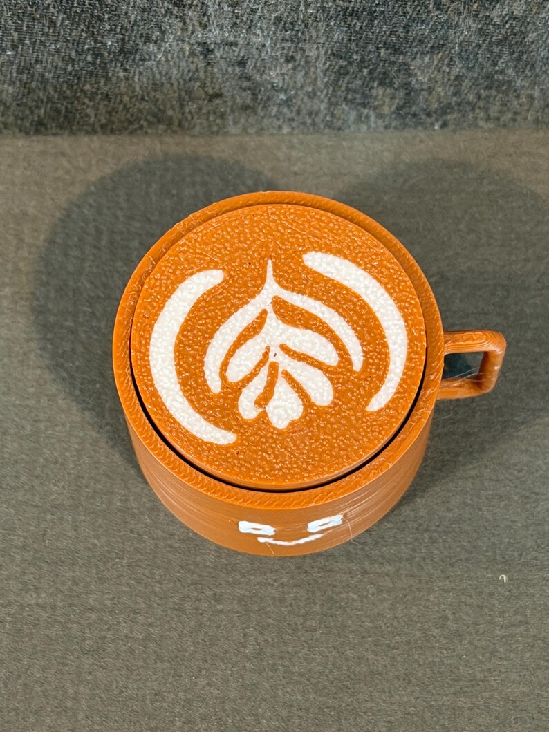 May include: A brown coffee cup with a white latte art design on top. The cup has a small handle and a smiling face design on the front. The cup is made of a solid material.