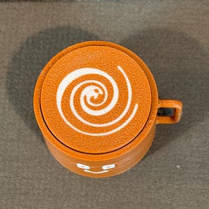 May include: An orange coffee cup with a white spiral design on top. The cup has a small handle and a smiling face design on the front. The cup is made of a solid material.