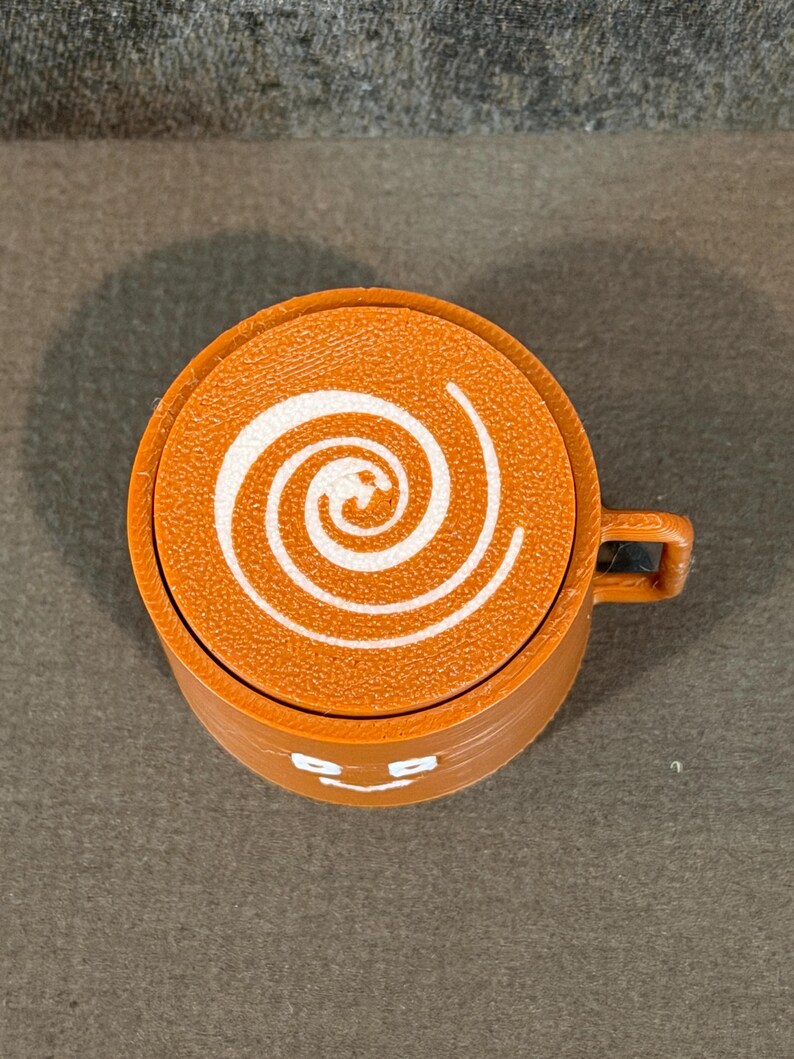 May include: An orange coffee cup with a white spiral design on top. The cup has a small handle and a smiling face design on the front. The cup is made of a solid material.