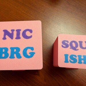 May include: Two pink square boxes with text. The larger box has the text "NIC" in purple and "BRG" in blue. The smaller box has the text "SQU" in purple and "ISH" in blue. Both boxes are on a wooden surface.