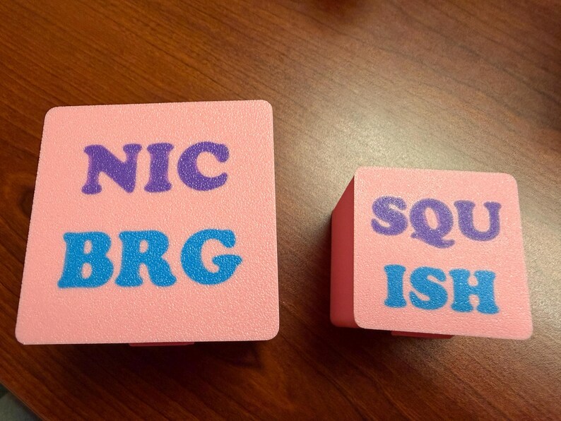 May include: Two pink square boxes with text. The larger box has the text "NIC" in purple and "BRG" in blue. The smaller box has the text "SQU" in purple and "ISH" in blue. Both boxes are on a wooden surface.