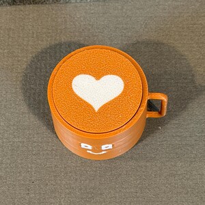May include: Orange coffee cup with a white heart design on the lid. The cup has a small handle and a smiling face design. The cup is made of a textured material.