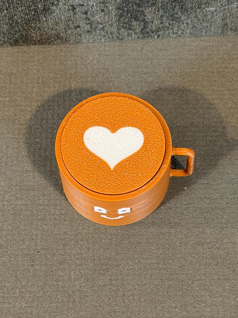 May include: Orange coffee cup with a white heart design on the lid. The cup has a small handle and a smiling face design. The cup is made of a textured material.