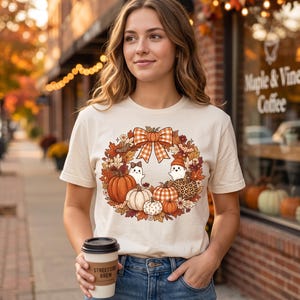 Peut inclure: Un t-shirt à manches courtes de couleur crème arborant un motif de couronne automnale composée de feuilles, de citrouilles et de deux petits fantômes. La couronne est surmontée d'un grand nœud à carreaux orange et blanc, avec des citrouilles à motifs.