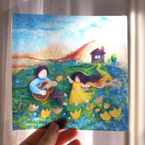 Op de afbeelding: Een naaldvilt kunstwerk met een persoon in een blauw shirt die op een bruine akoestische gitaar speelt, terwijl een ander persoon in een gele jurk in de buurt danst. De scène toont een groene weide, gele bloemen en een klein bruin huisje.