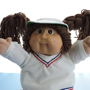 May include: A soft-sculpture doll with brown yarn hair styled in two high pigtails. The doll wears a white long-sleeved shirt with a V-neck featuring red and blue trim, and a white cap with a green brim. The doll has a round face with dark eyes.