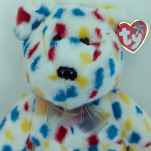 May include: A white plush toy bear with a colorful confetti pattern. The bear has a red, yellow, and blue confetti pattern. The bear has a red Ty tag with a white star.