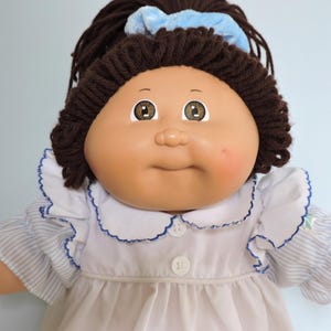 May include: A doll with dark brown yarn hair styled in a ponytail with a light blue scrunchie. The doll has a round face, brown eyes, and a light pink cheek blush. It wears a white dress with a blue-trimmed collar and blue-and-white striped sleeves.