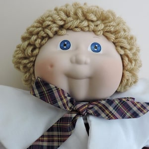 May include: A doll with a round, light-toned face, bright blue eyes, and a single dimple on the left cheek. The doll has thick, curly blonde yarn hair and wears a white collared shirt accented with a dark plaid necktie featuring red and yellow lines.